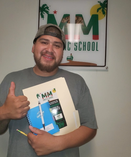 Man in gray shirt holding notebook and folder, giving thumbs up, smiling near a school sign.