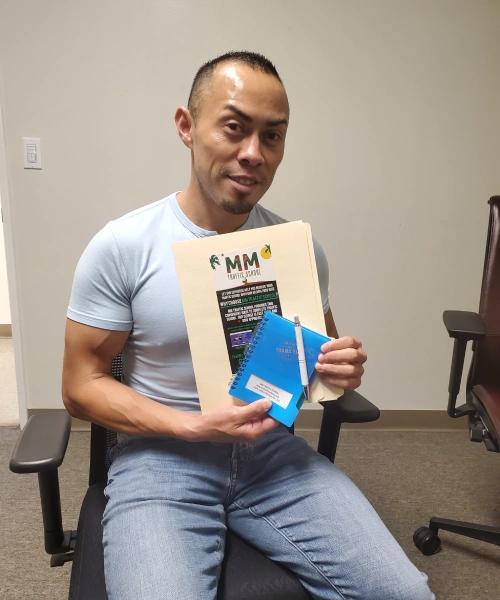 Man holding a folder and notebook, smiling. He's wearing a blue shirt and jeans, seated indoors near a desk.