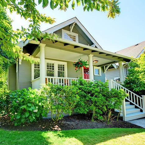 A house with a large porch and stairs is surrounded by trees and bushes.