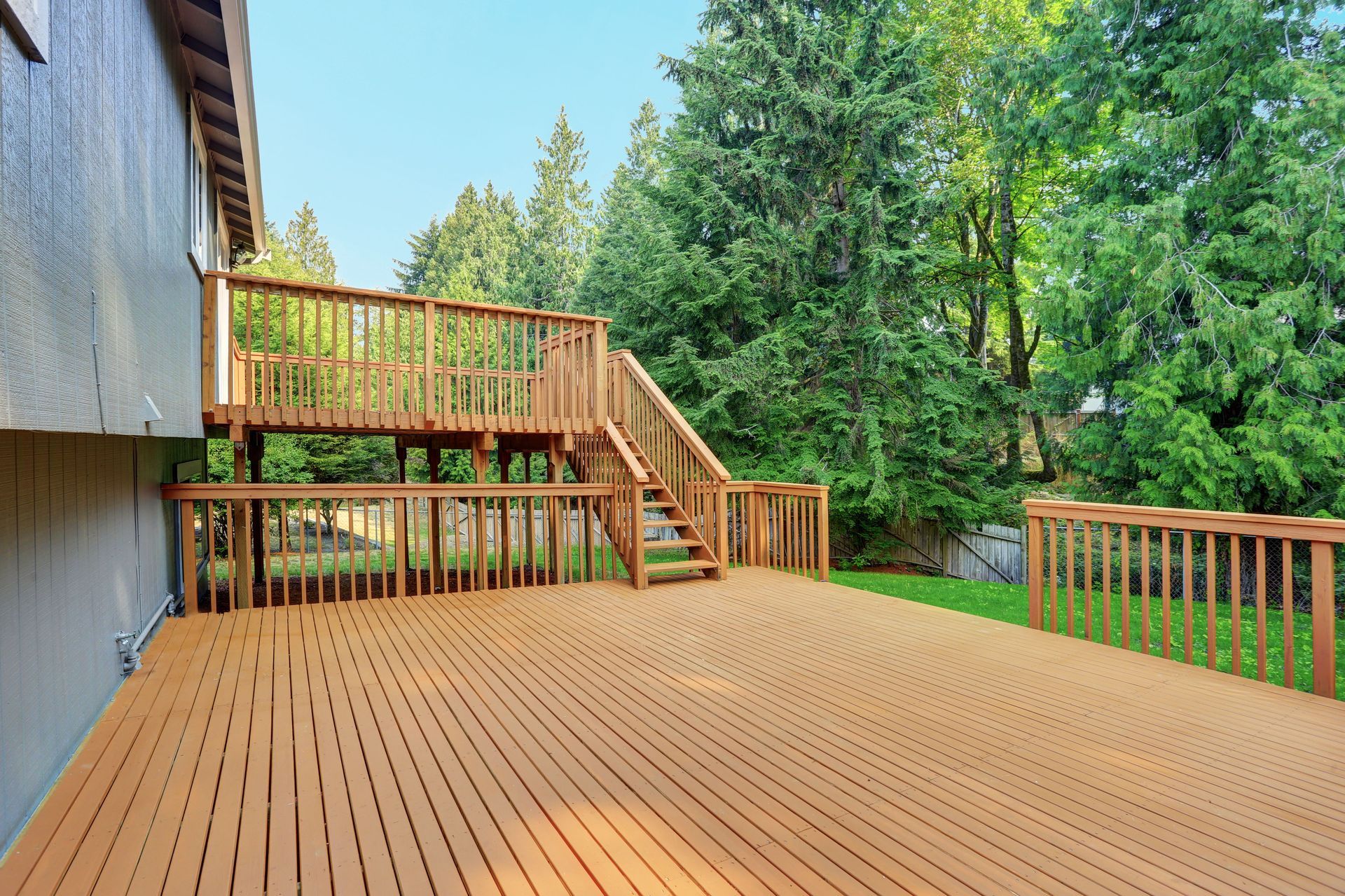 Wooden two-level deck attached to a gray house, stairs leading up, surrounded by trees.