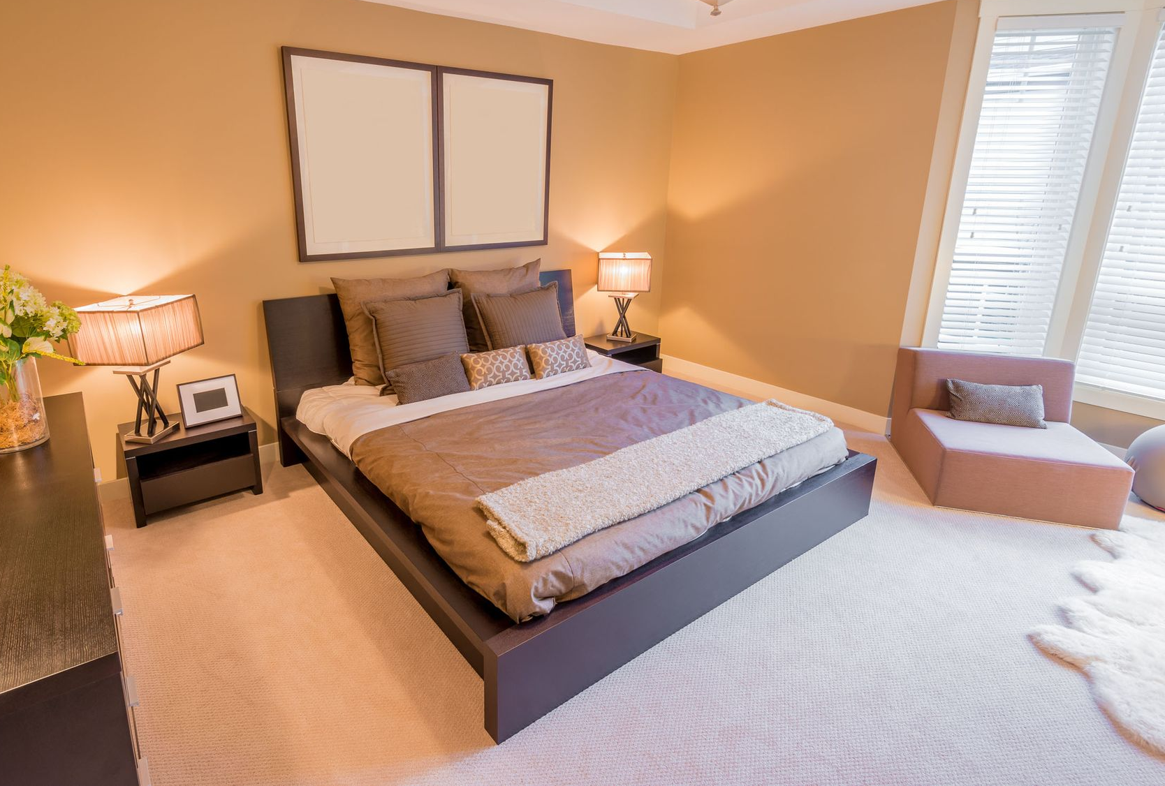 Bedroom with a bed, nightstands with lamps, a small chair, and a large window. Cream and brown tones.