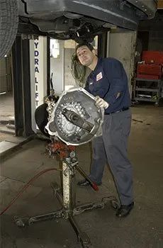 Mechanic standing beside a lifted car, inspecting a transmission on a stand in a garage.