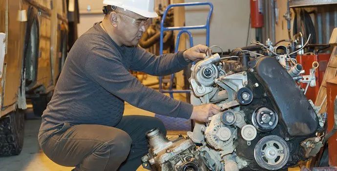 Mechanic kneeling beside a car engine, tightening parts in a workshop with tools and equipment nearby