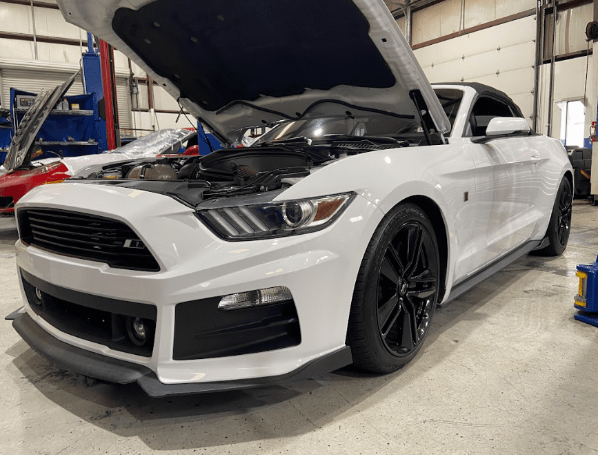 A white ford mustang with the hood up is parked in a garage.