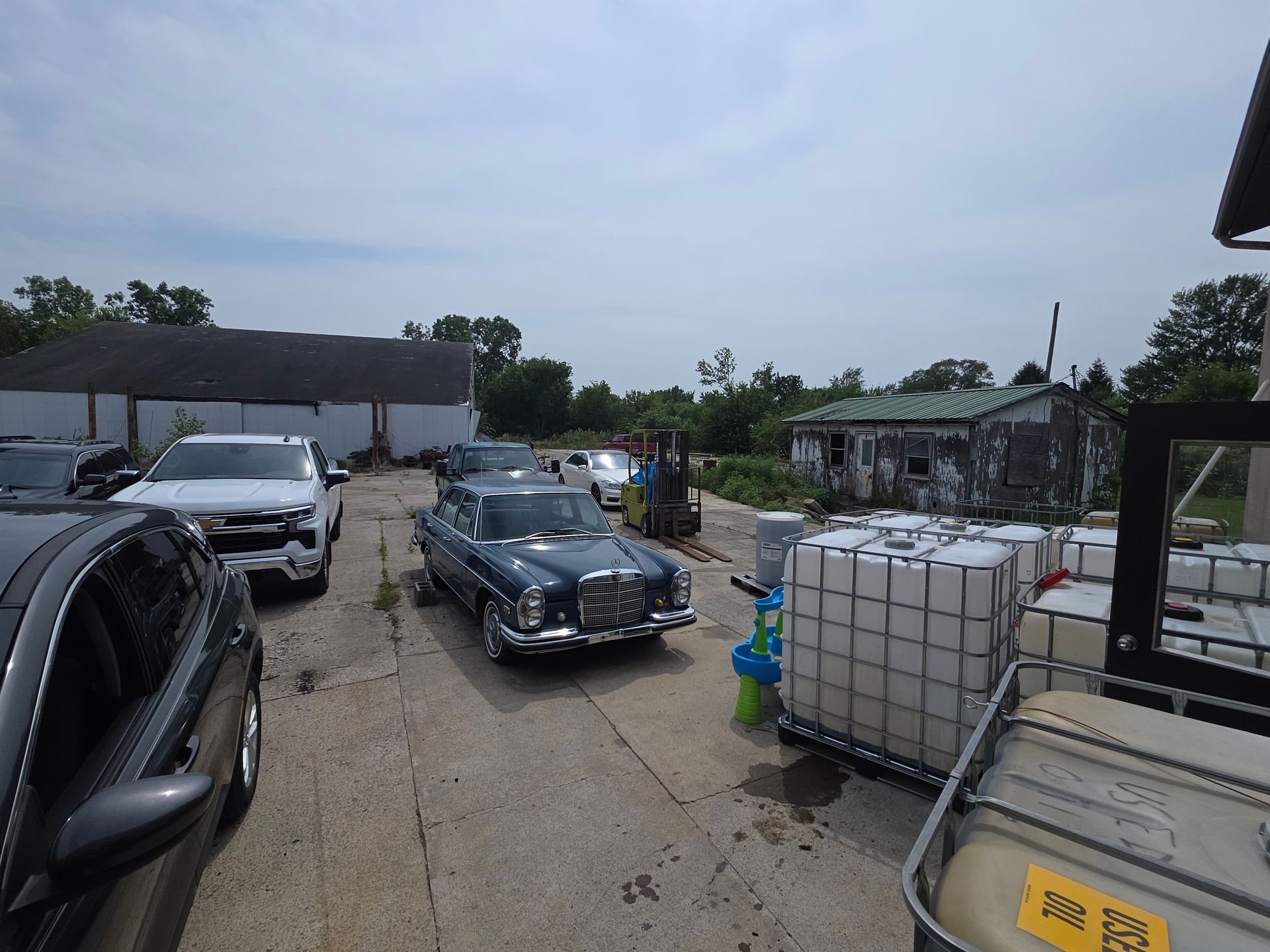 Classic cars parked on a wet lot beside stacked water containers under a cloudy sky.