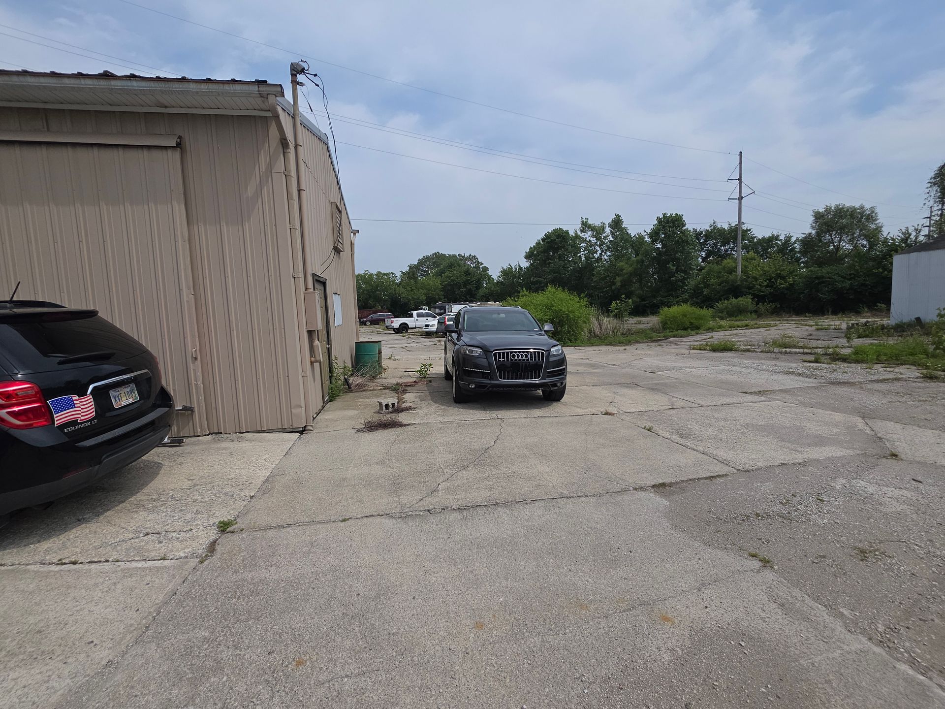 Two cars parked by a tan building on a cracked, empty lot under a cloudy sky