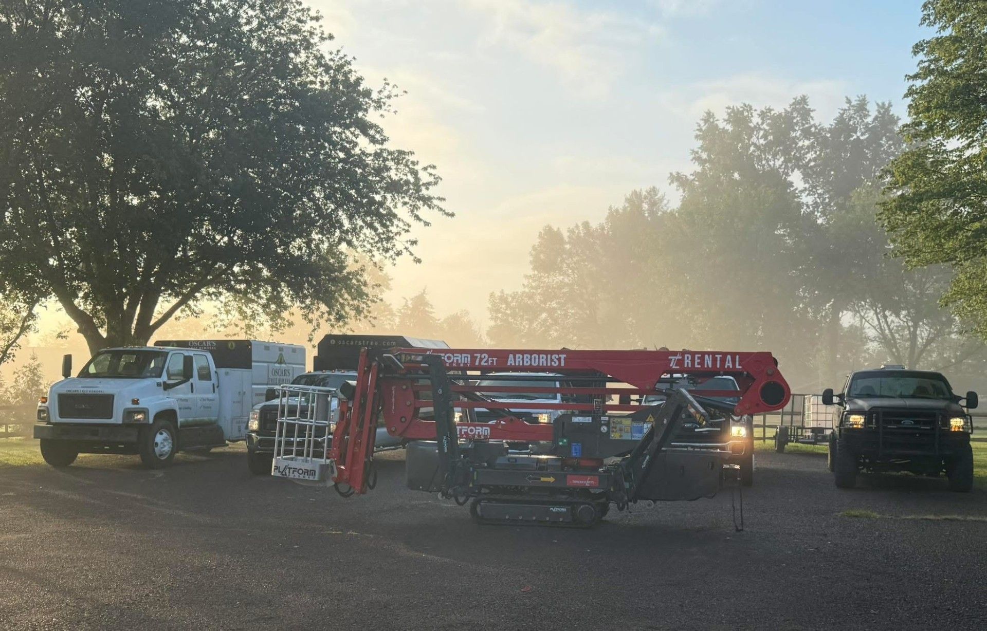Vehicles parked outside on a foggy day. A red lift and two trucks sit on gravel with trees in the background.