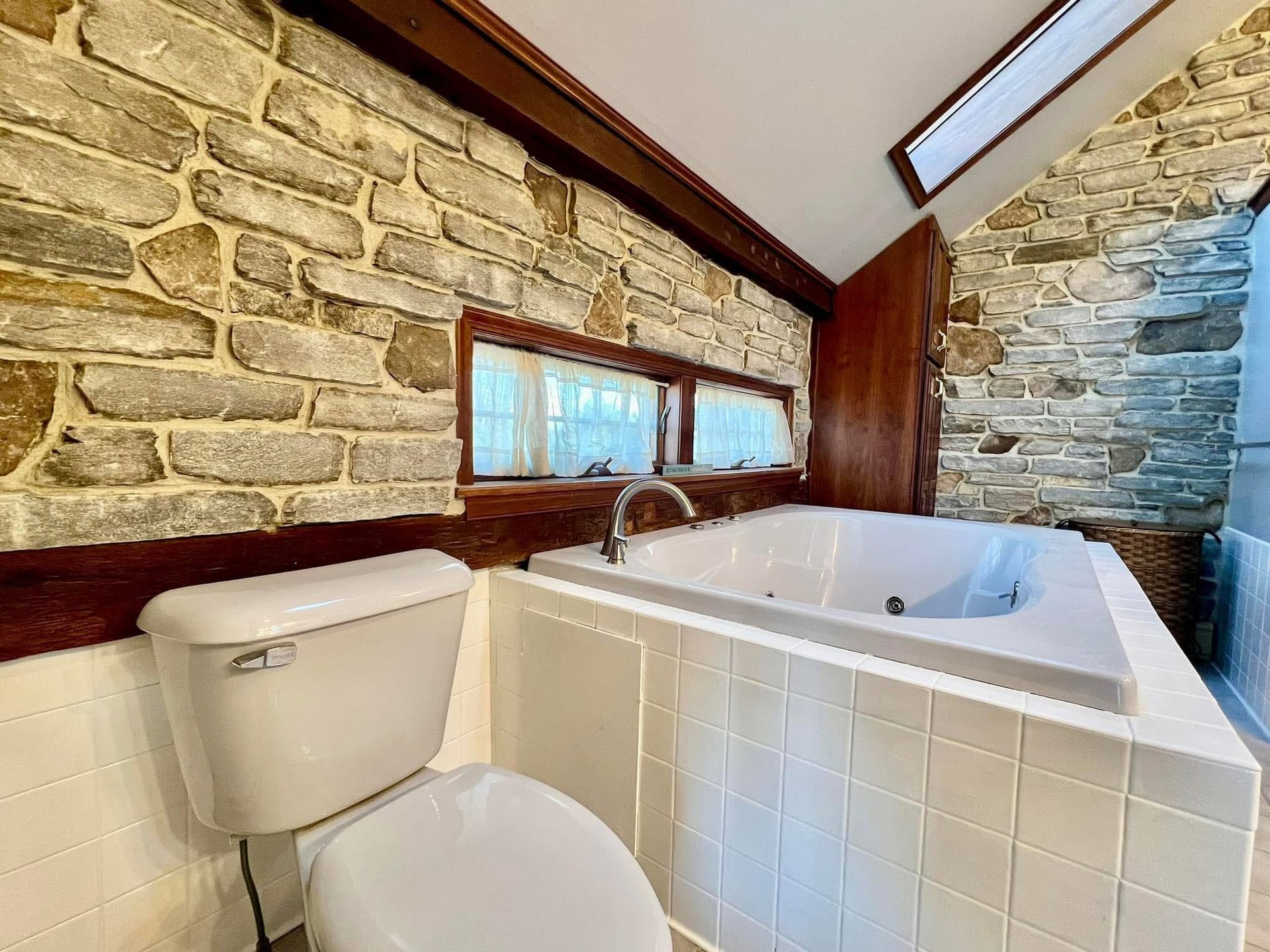 A bathroom with a toilet , jacuzzi tub , and stone wall.