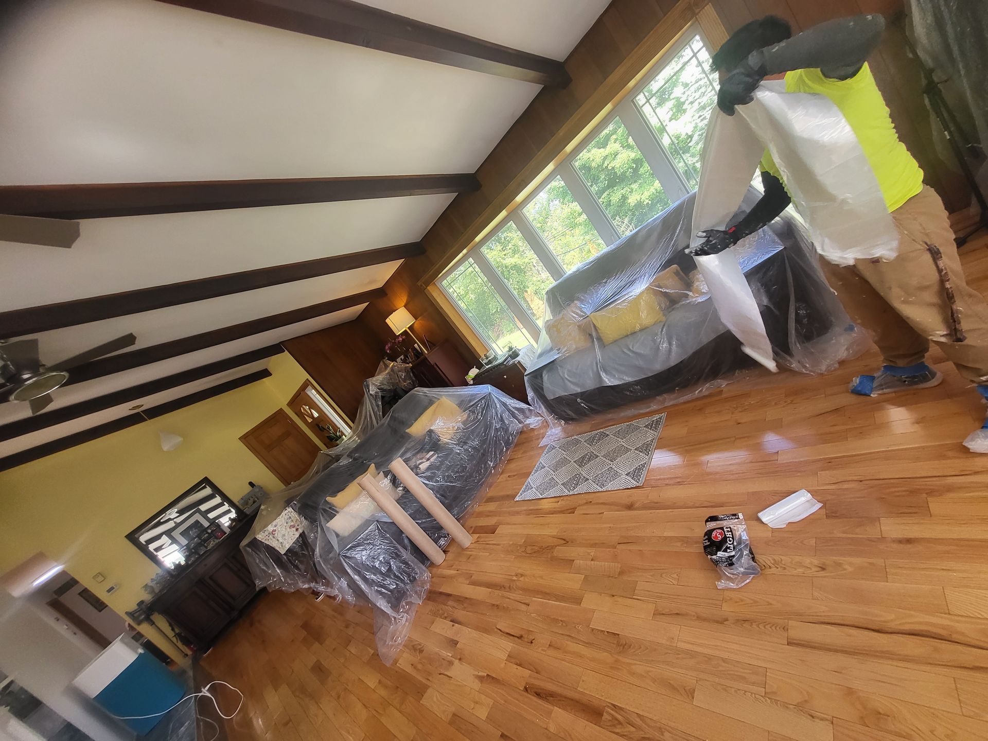 a man in a yellow-green shirt is standing in a living room covering a couch with plastic wrap