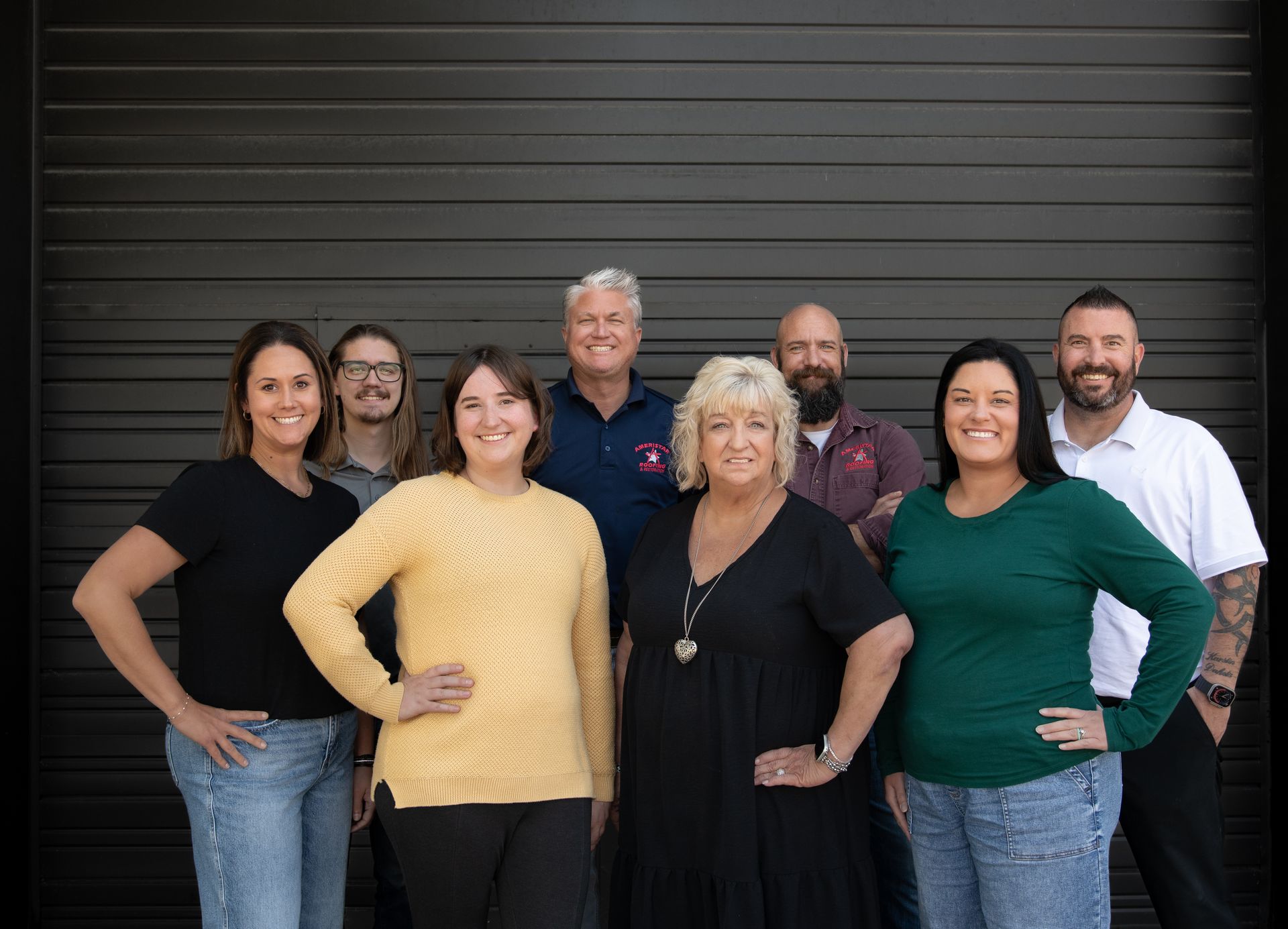 Eight people posing together in front of a dark garage door, smiling at the camera.