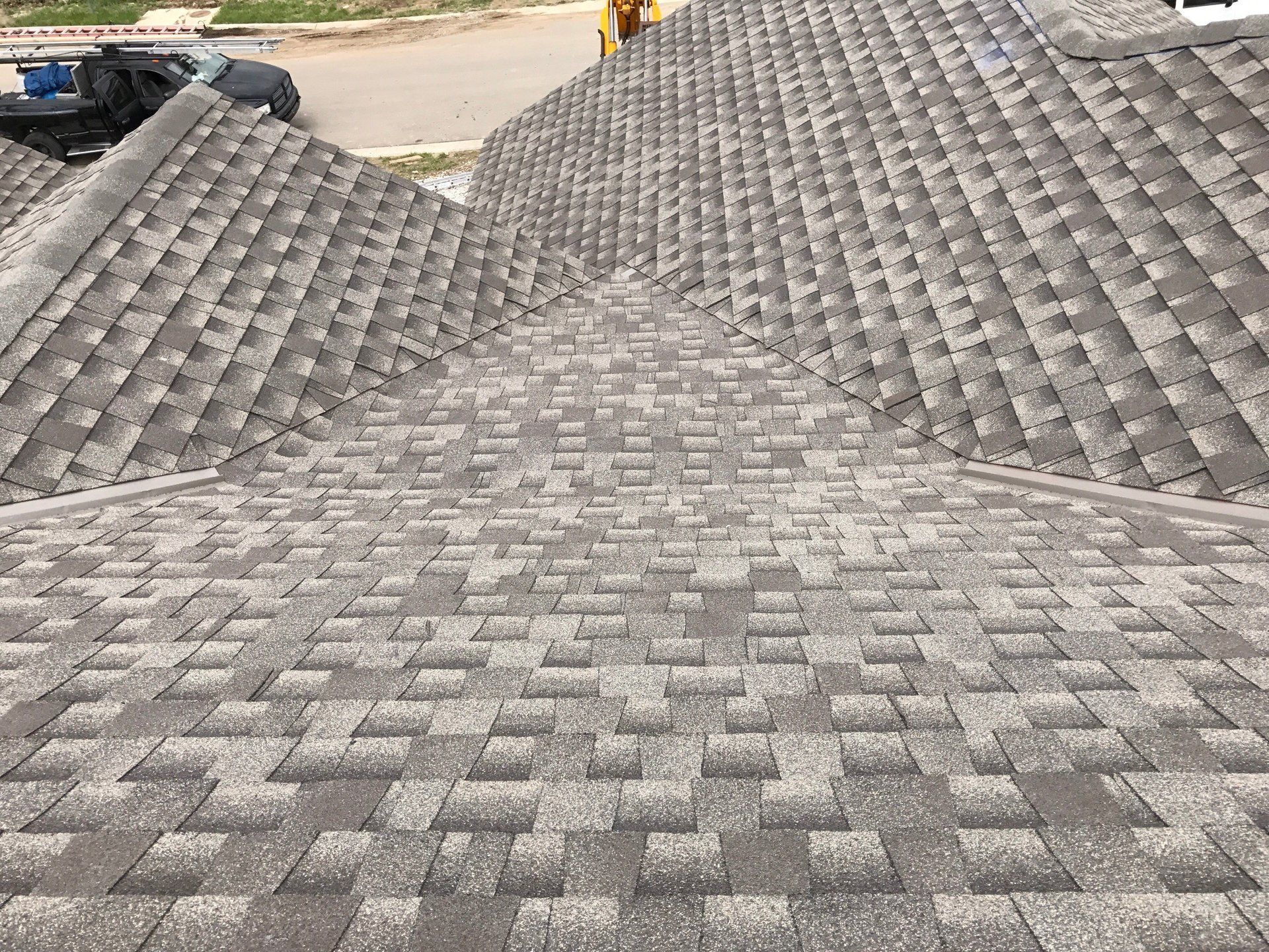A top-down view of a grey shingled roof with multiple sloped sections meeting at a central valley near a street.