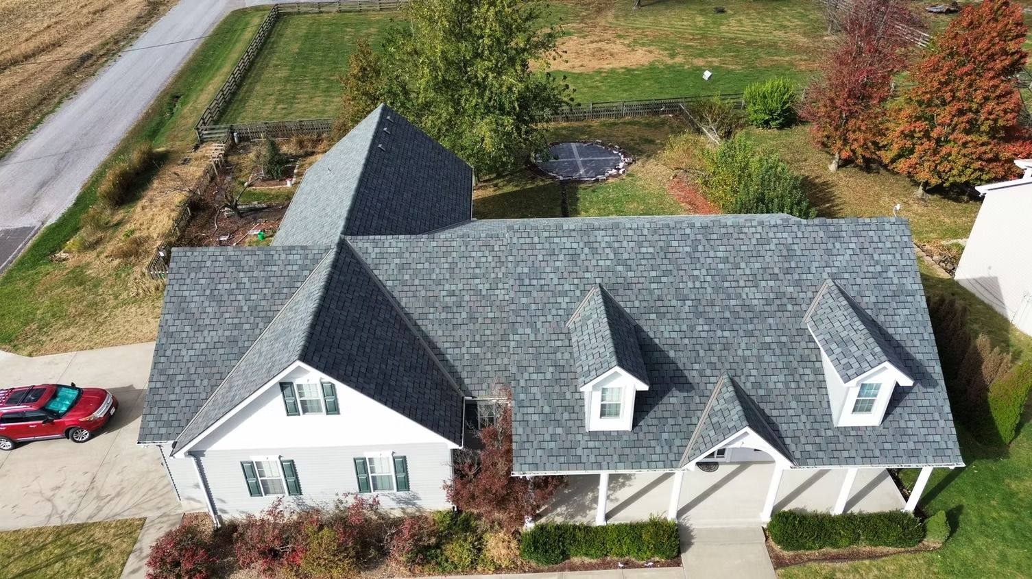 Aerial view of a house with a gray shingled roof, two dormers, and a white exterior, located on a grassy suburban lot.