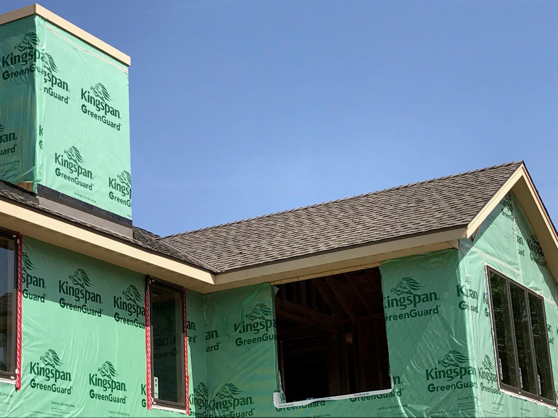 A close-up of a house under construction, featuring green protective sheeting and newly installed roof shingles.