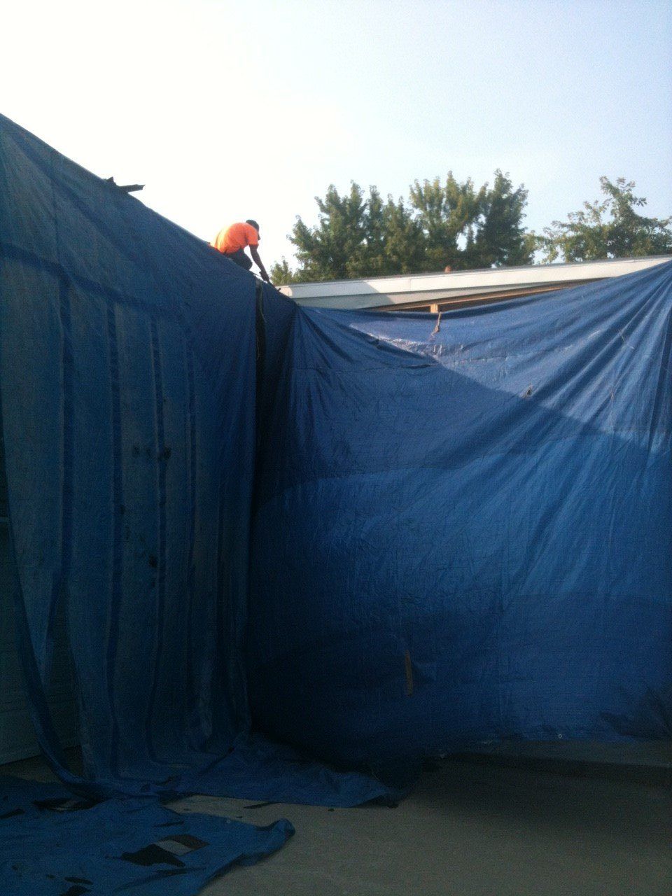 A worker in an orange shirt stands on a roof covered by a large, blue tarp draped over the side of a building.