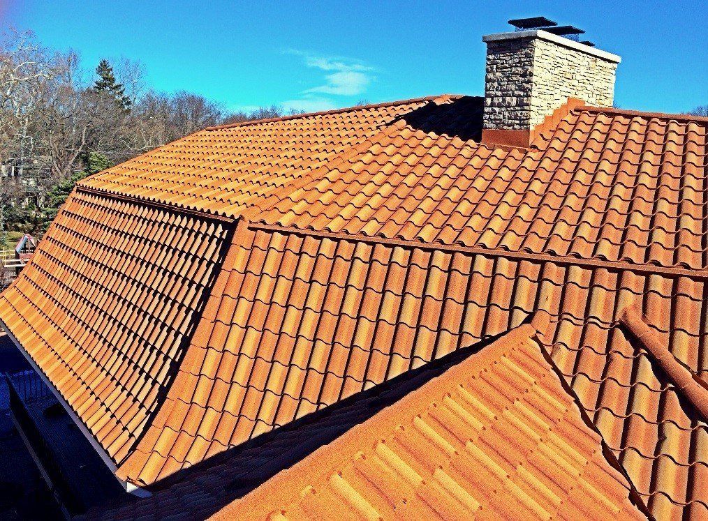 A high-angle view of a terracotta tiled roof under a bright blue sky, featuring a stone chimney.