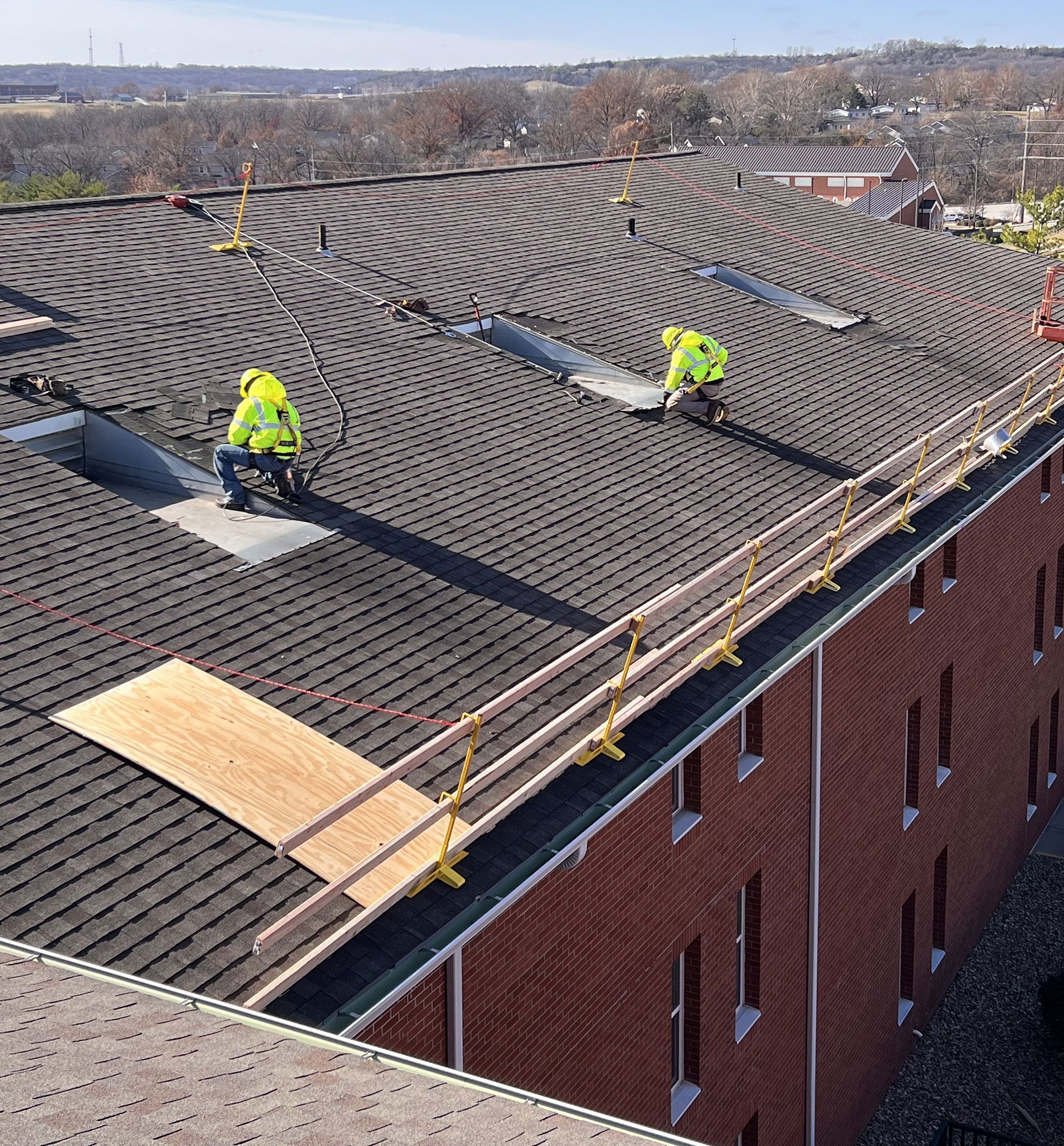 Two workers in neon safety gear install a patterned dark shingle roof on a brick building with a safety railing.