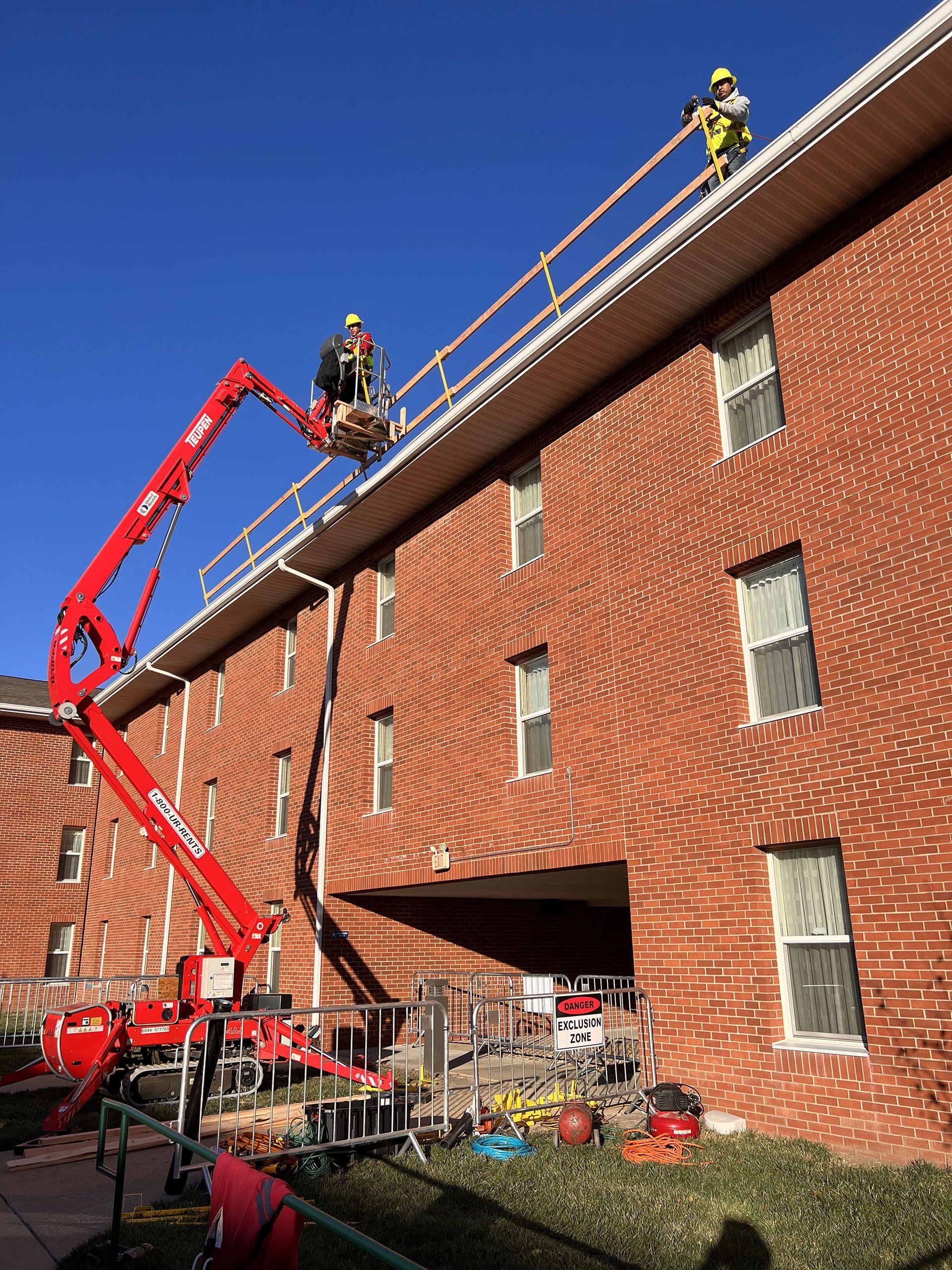 Workers in a red articulating boom lift repair the roof of a red brick building under a clear blue sky.