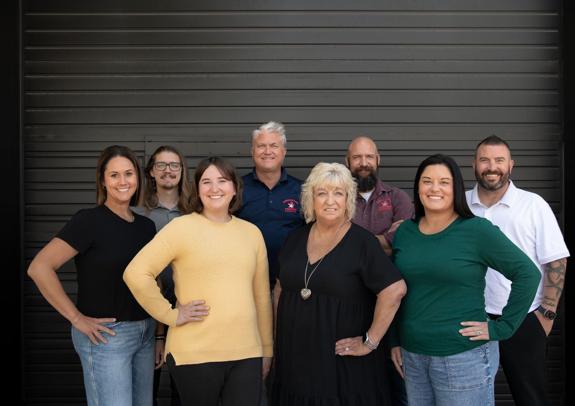 Seven people posing in front of a dark gray metal wall, smiling at the camera.