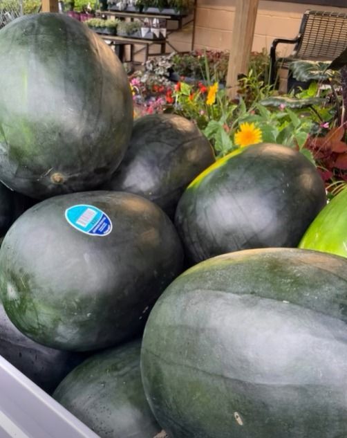 Large dark green watermelons stacked at a market stand with flowers in the background