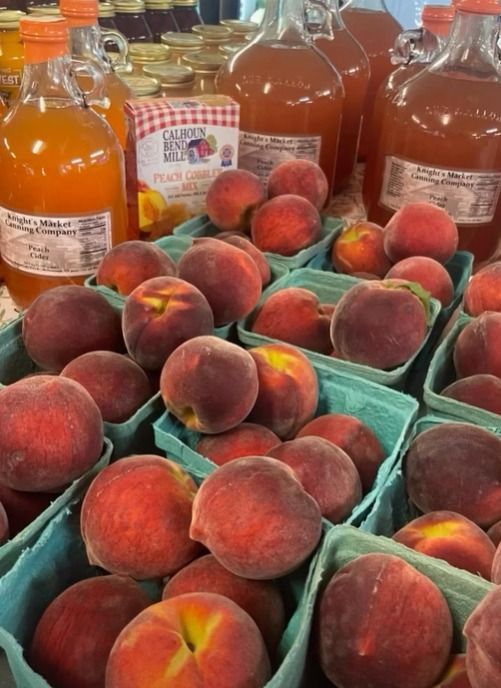 Peaches piled in green crates at a market, with bottles of juice behind them.