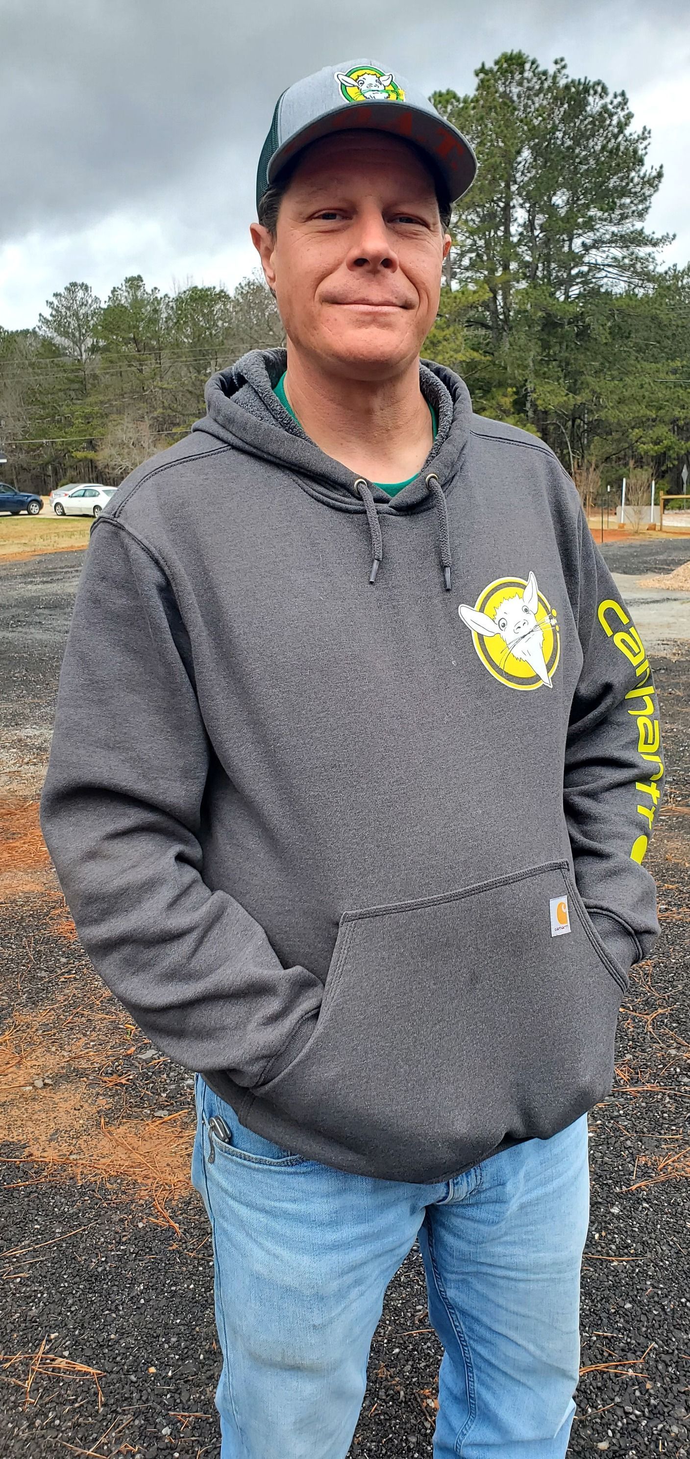 A man wearing a hoodie and a hat is standing in a gravel lot.
