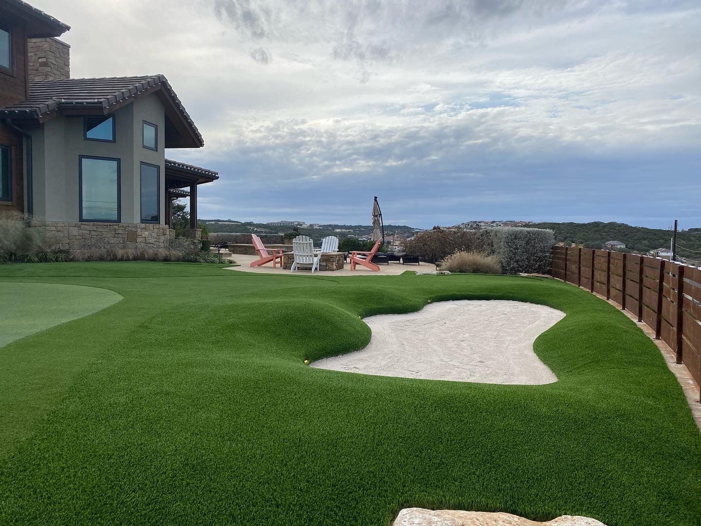 Backyard with a golf green, sand trap, seating area, and a house overlooking a cloudy landscape.