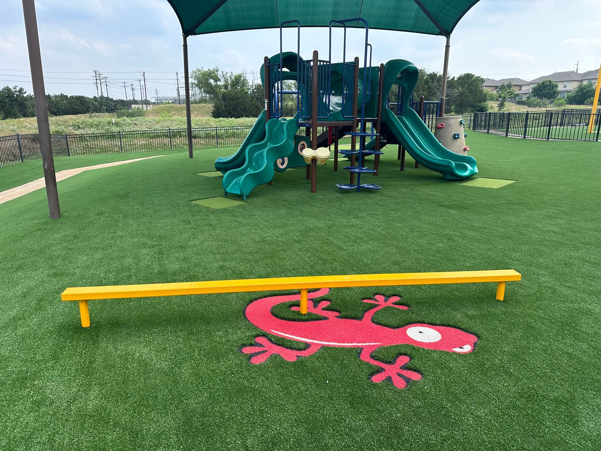 Playground with green slides, a yellow balance beam over a red lizard on green turf, under a canopy.