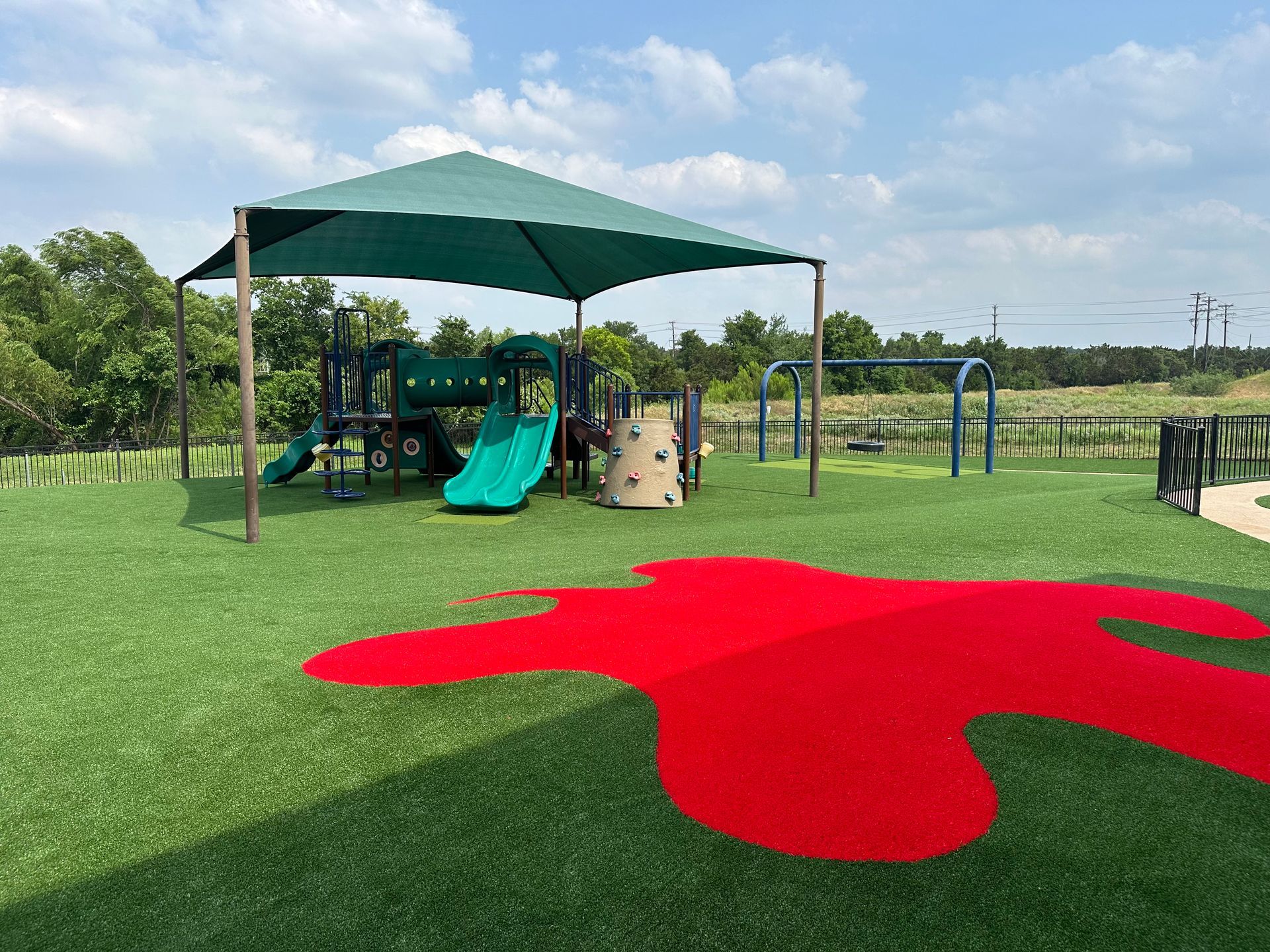 Playground with green turf, a red splash, and a shaded area with slides, climbing structures, and swings.