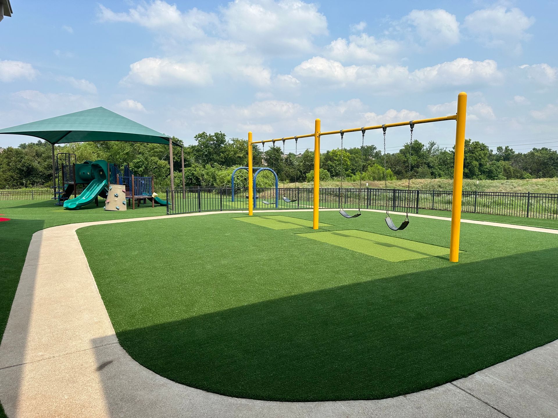 Playground with yellow swings, green turf, and shade structure, outdoors on a sunny day.