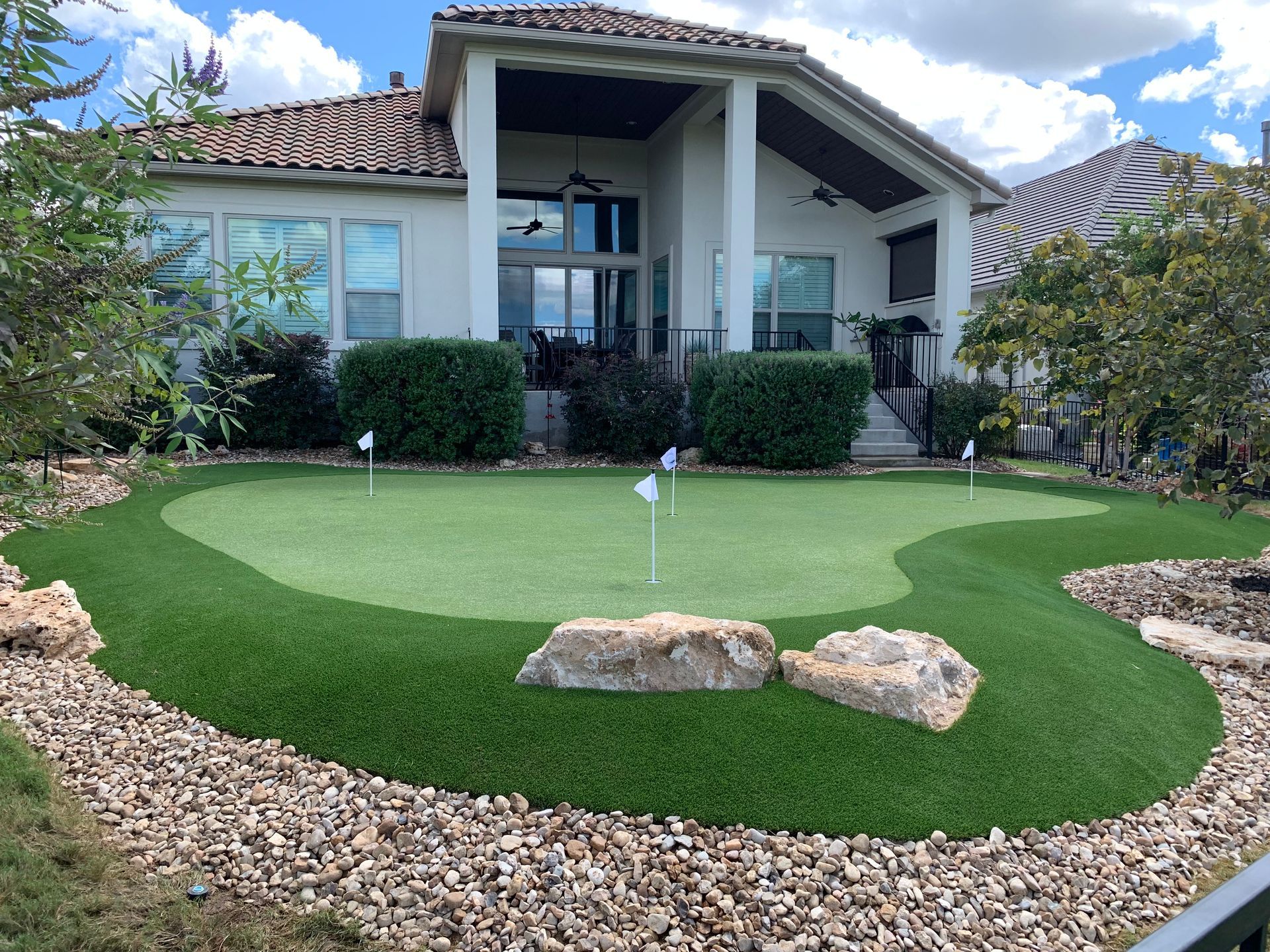Backyard putting green with flags, rocks, and house in the background. Green turf, blue sky.
