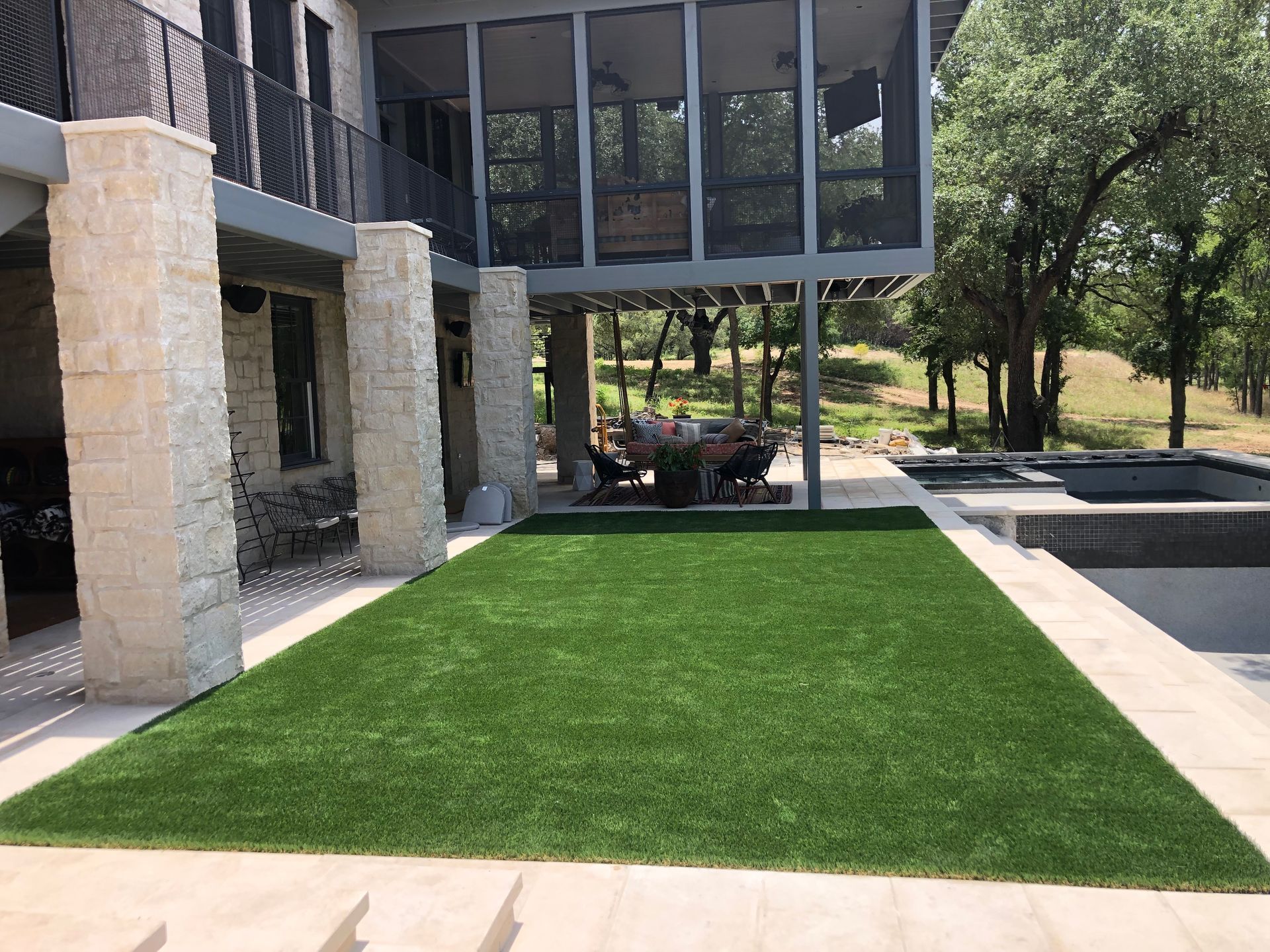 Green lawn next to a patio and pool, under a house with stone columns. People seated at a table.
