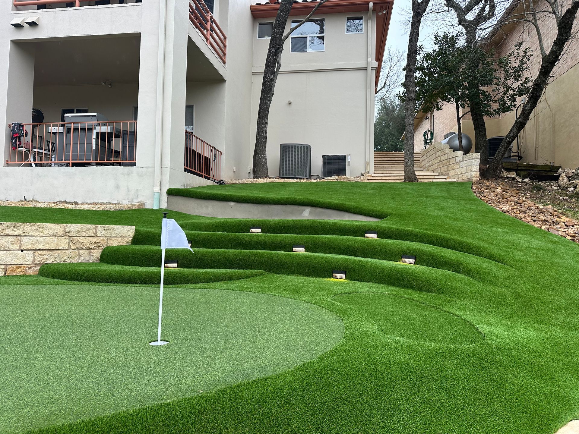 A backyard putting green with tiered artificial turf levels. A white flag marks the hole.