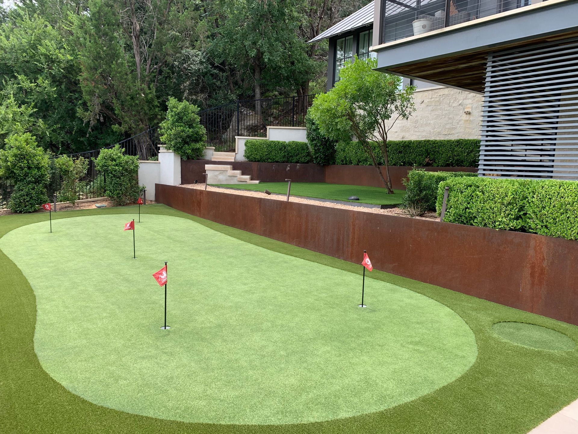 A putting green with red flags, bordered by a rust-colored wall and green hedges, next to a modern building.