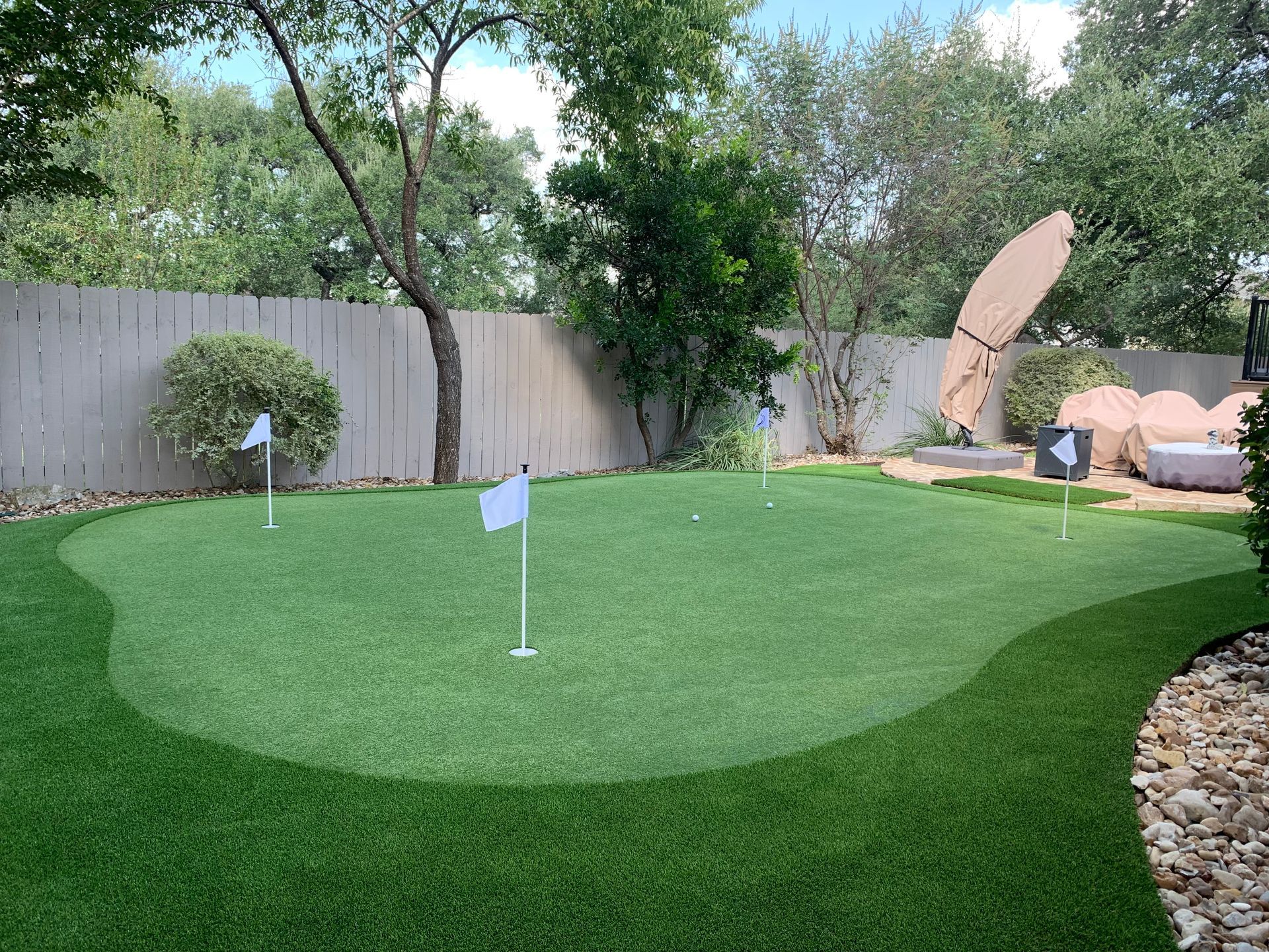 Green artificial turf putting green with four holes, white flags, and a wooden fence.