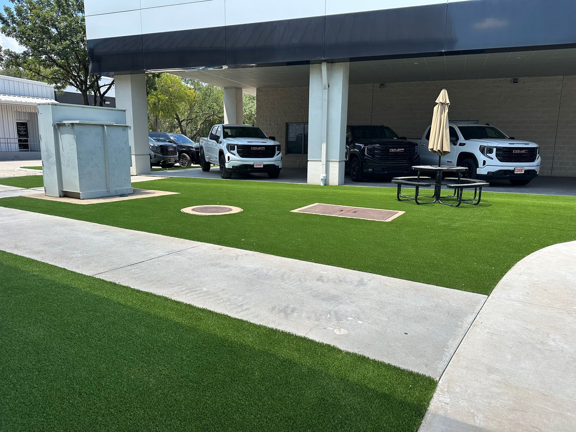 Several GMC trucks parked under a building overhang with picnic tables on green turf.