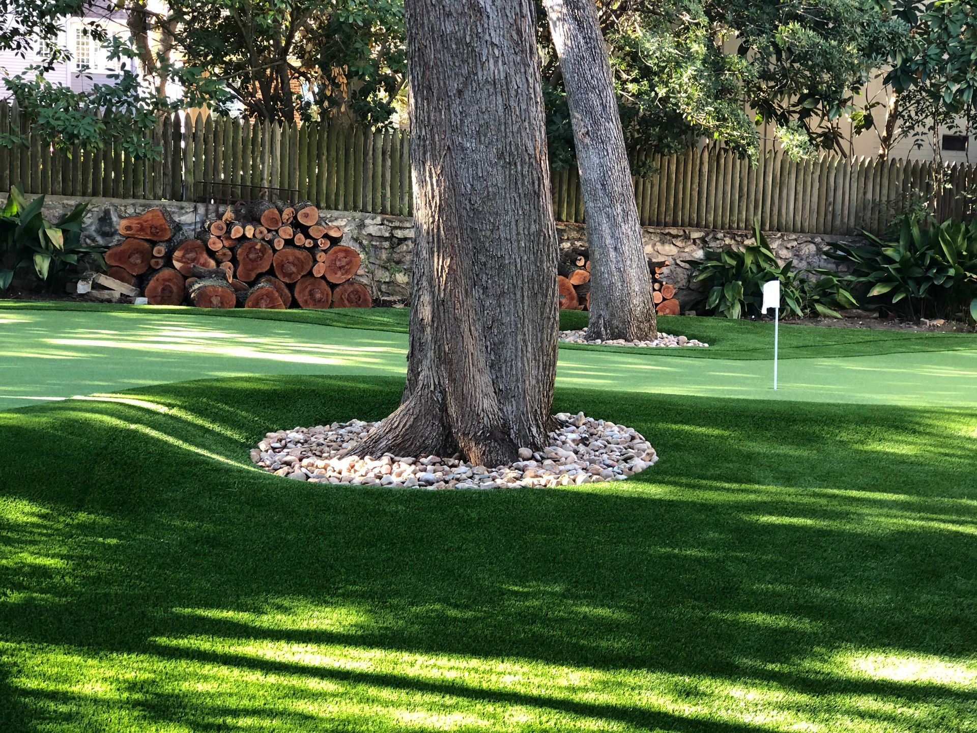 A putting green with trees, featuring a golf hole and a pile of firewood in the background.