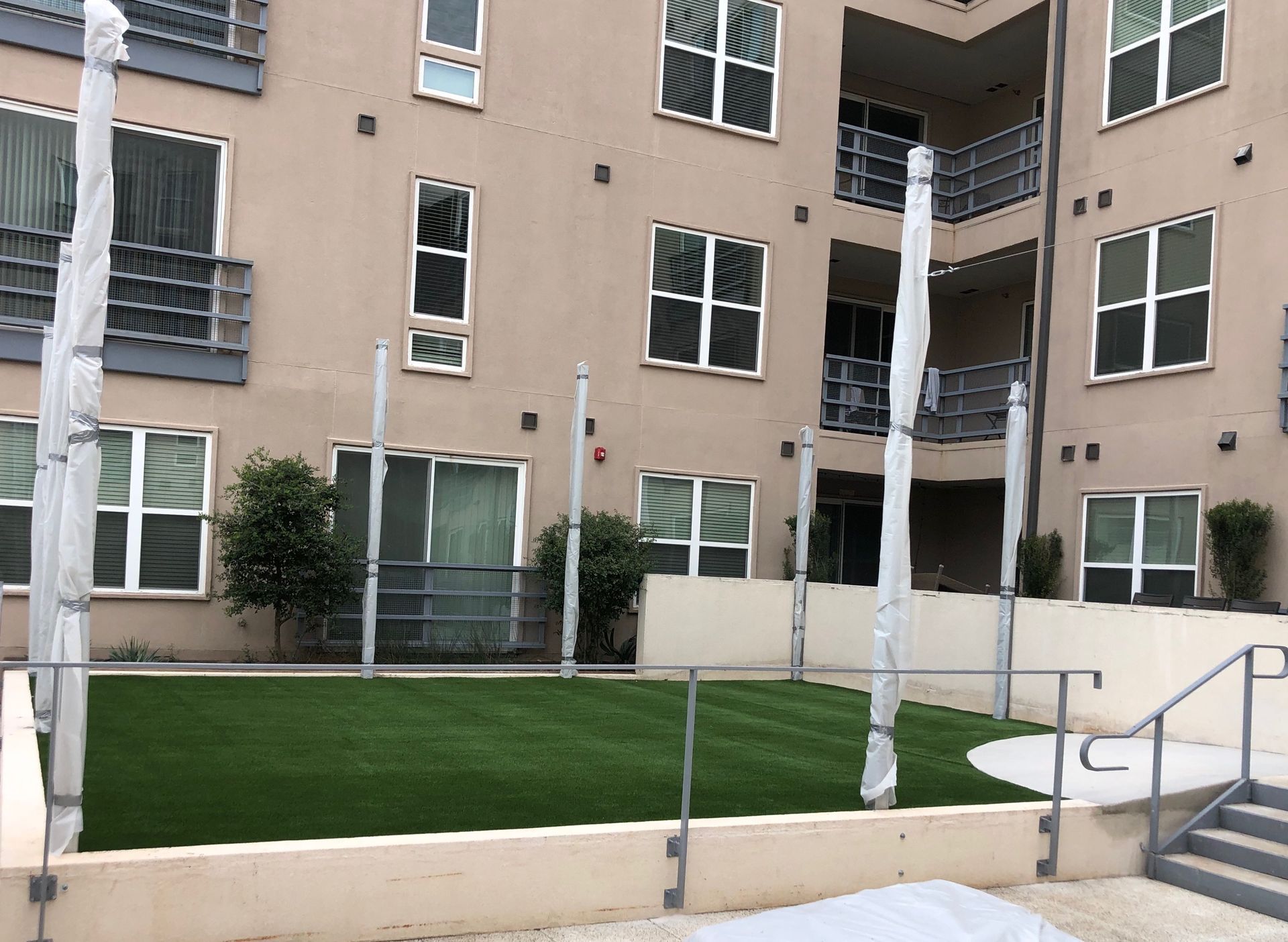 Courtyard with green turf surrounded by a low wall, building in the background. Columns wrapped in plastic.