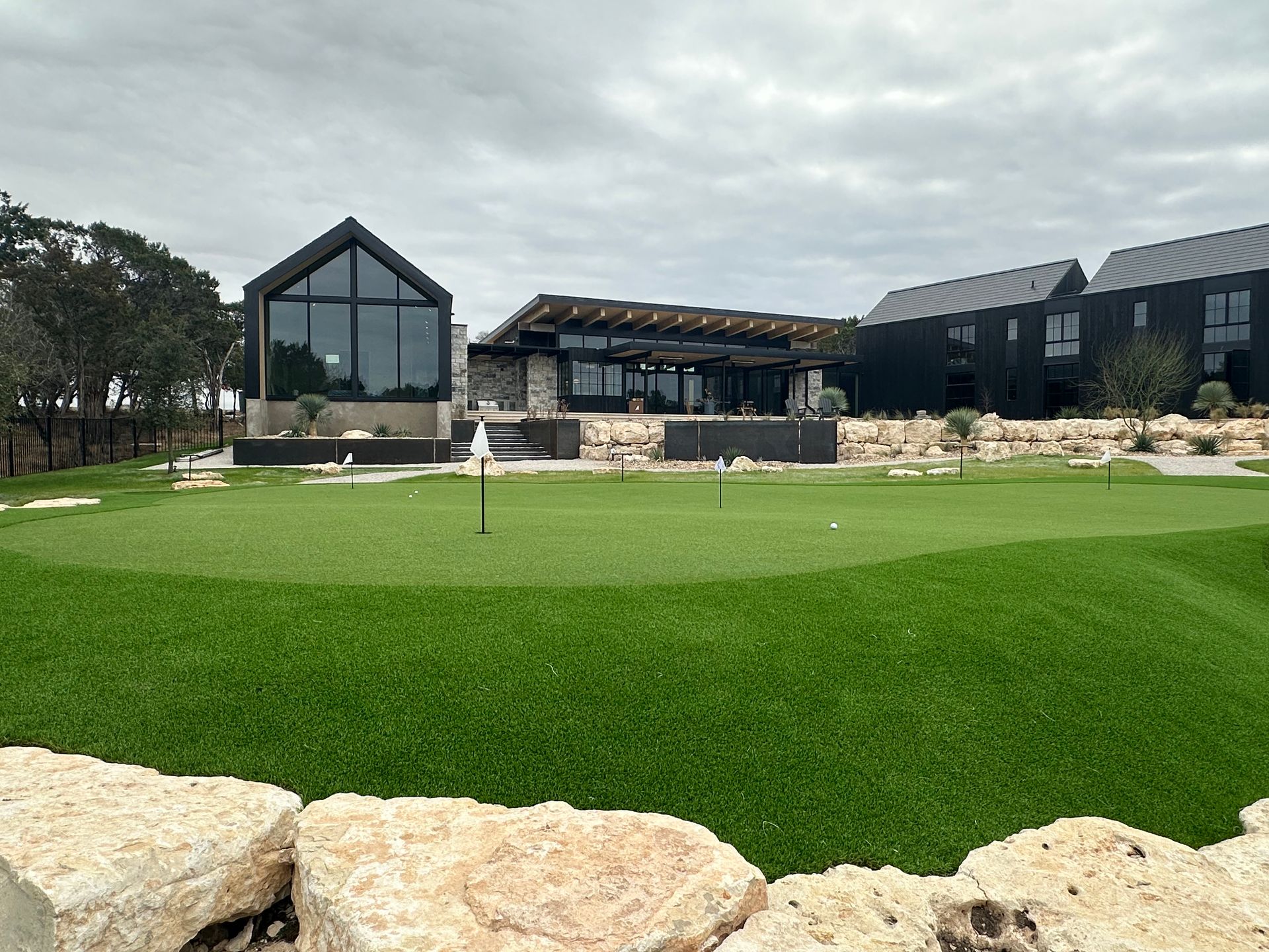 A modern putting green with flagsticks, stone accents, and dark buildings under a cloudy sky.