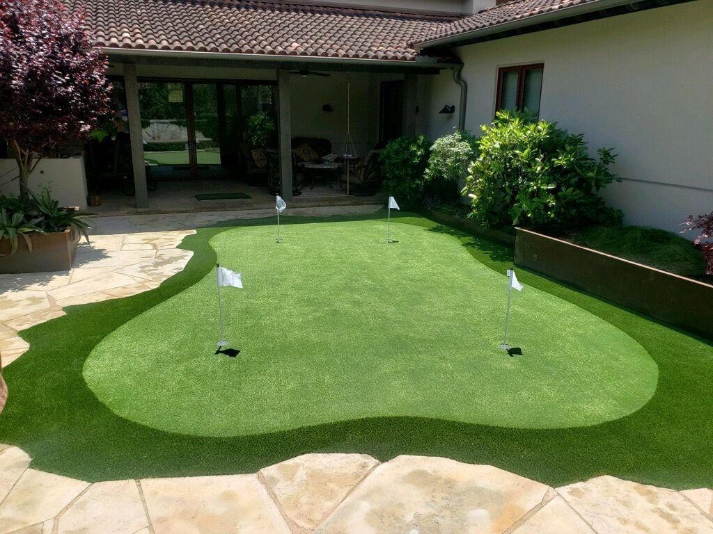 Artificial turf putting green in a backyard with flagsticks, surrounded by stone paving and landscaping.