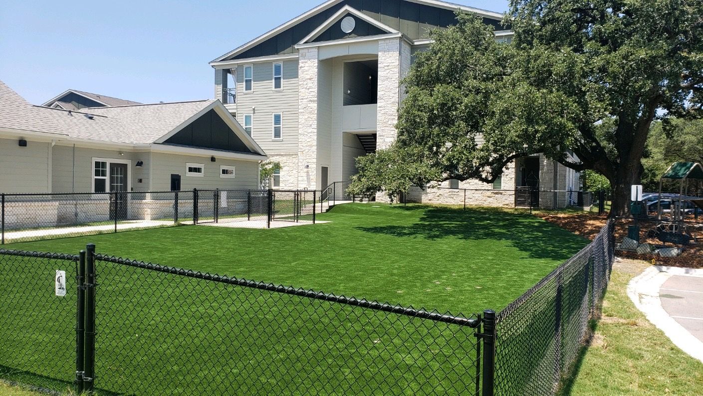 Fenced dog park with artificial turf in front of a multi-story apartment building on a sunny day.