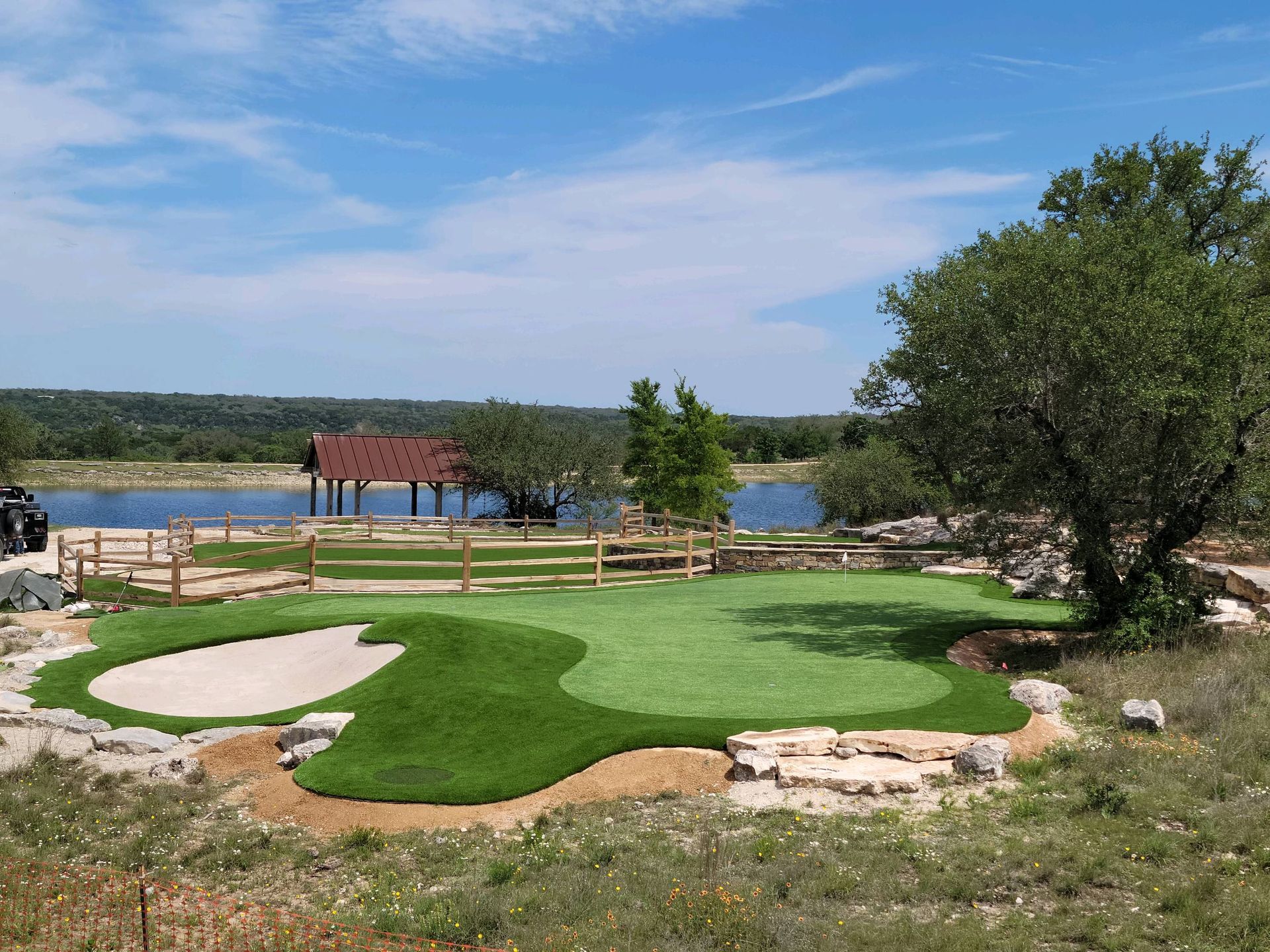 A putting green with sand trap, wooden fence, lake, and small pavilion on a sunny day.