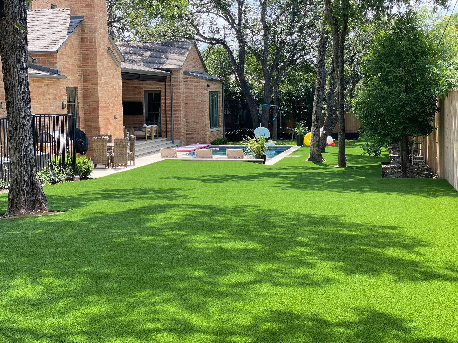 Backyard with green artificial grass, pool, brick house, and trees.