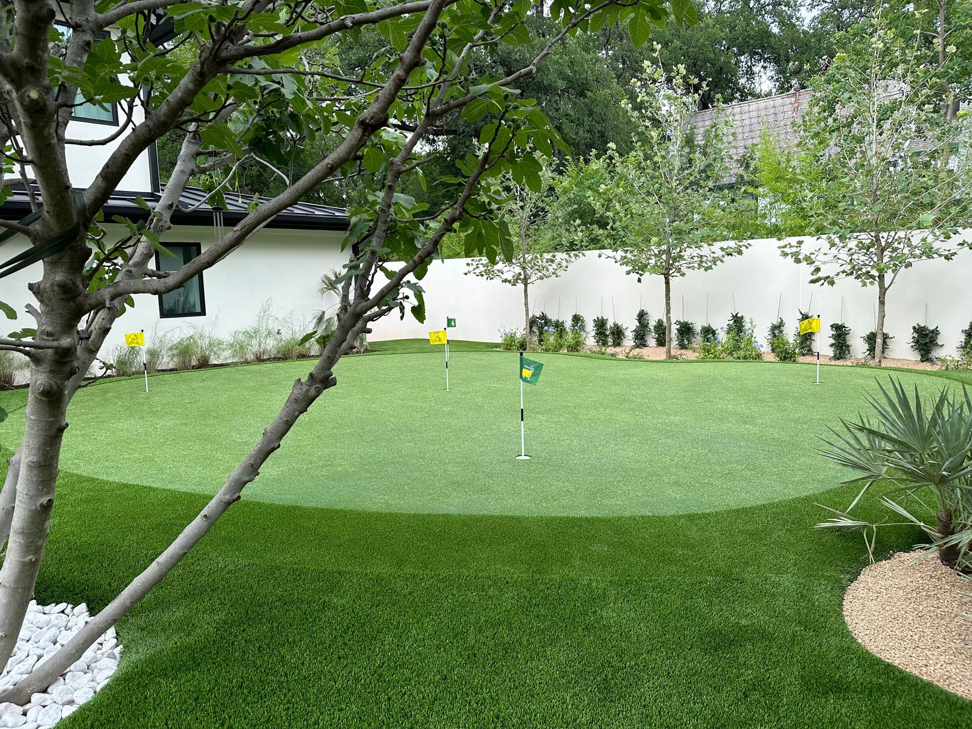 A putting green with three holes and flags, surrounded by white fence and trees.