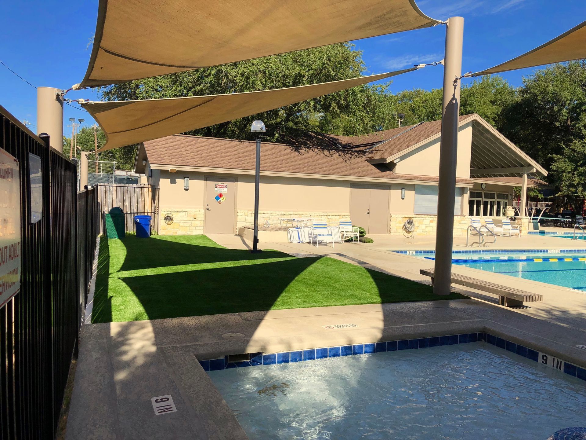 Poolside area with shade sails, a building, and a small pool. Green grass and blue water.