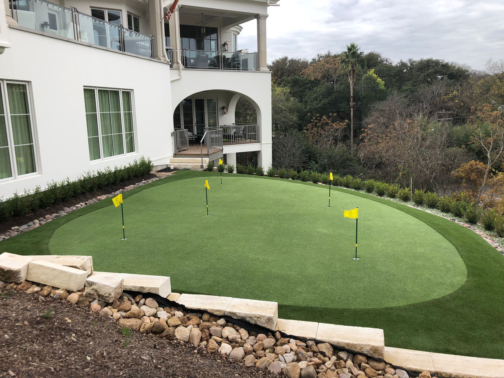 A backyard putting green with flags, next to a white house with a balcony.
