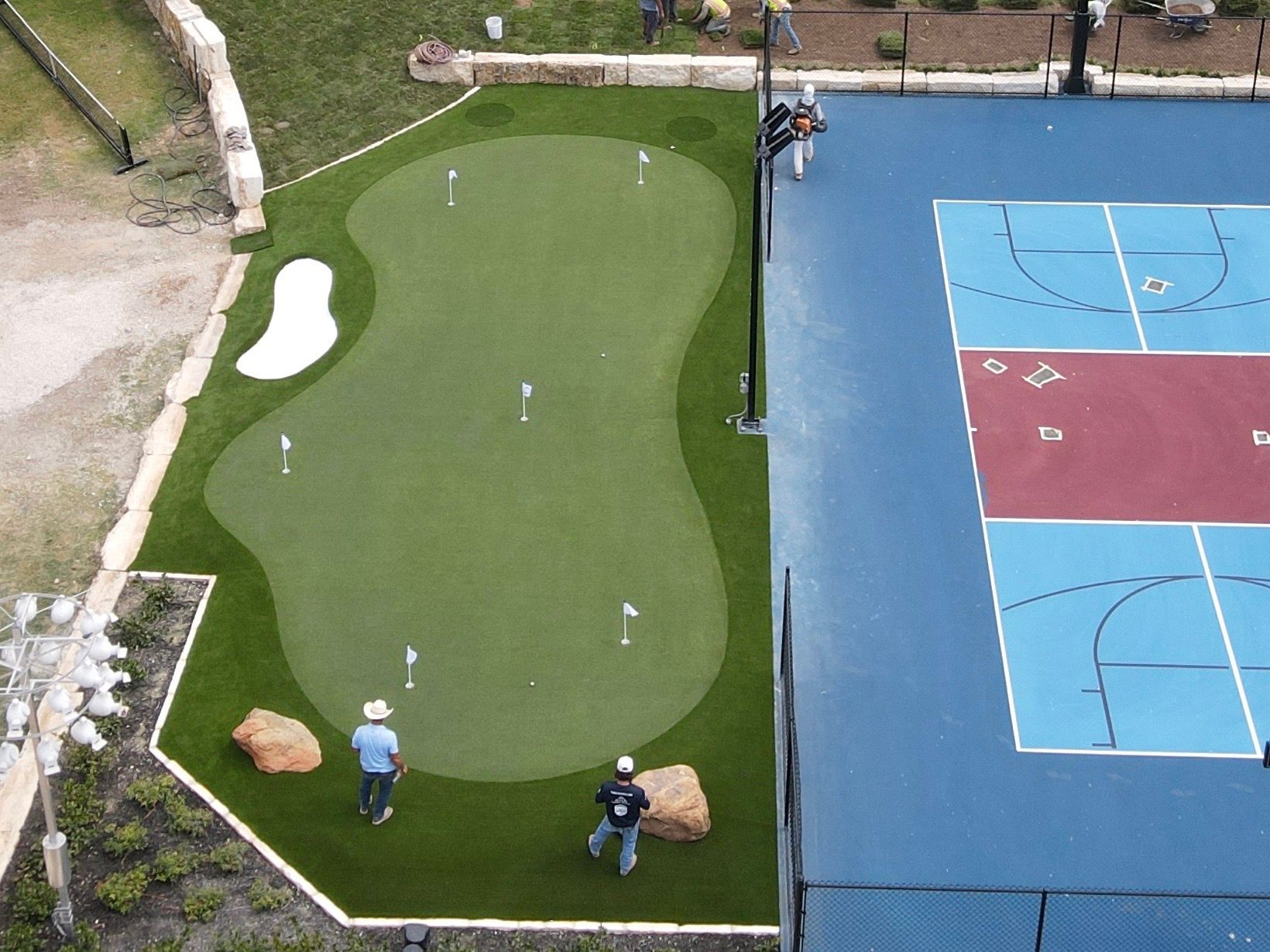 Aerial view of a green putting green next to a blue basketball court. Two men are on the green, practicing golf.