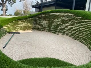 Golf course sand trap with raked sand. Green grass surrounds the trap, with a rake resting inside.