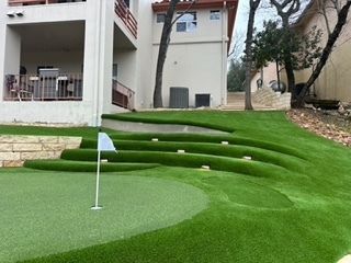Backyard putting green with tiered turf seating, flagstick, and multi-story house.
