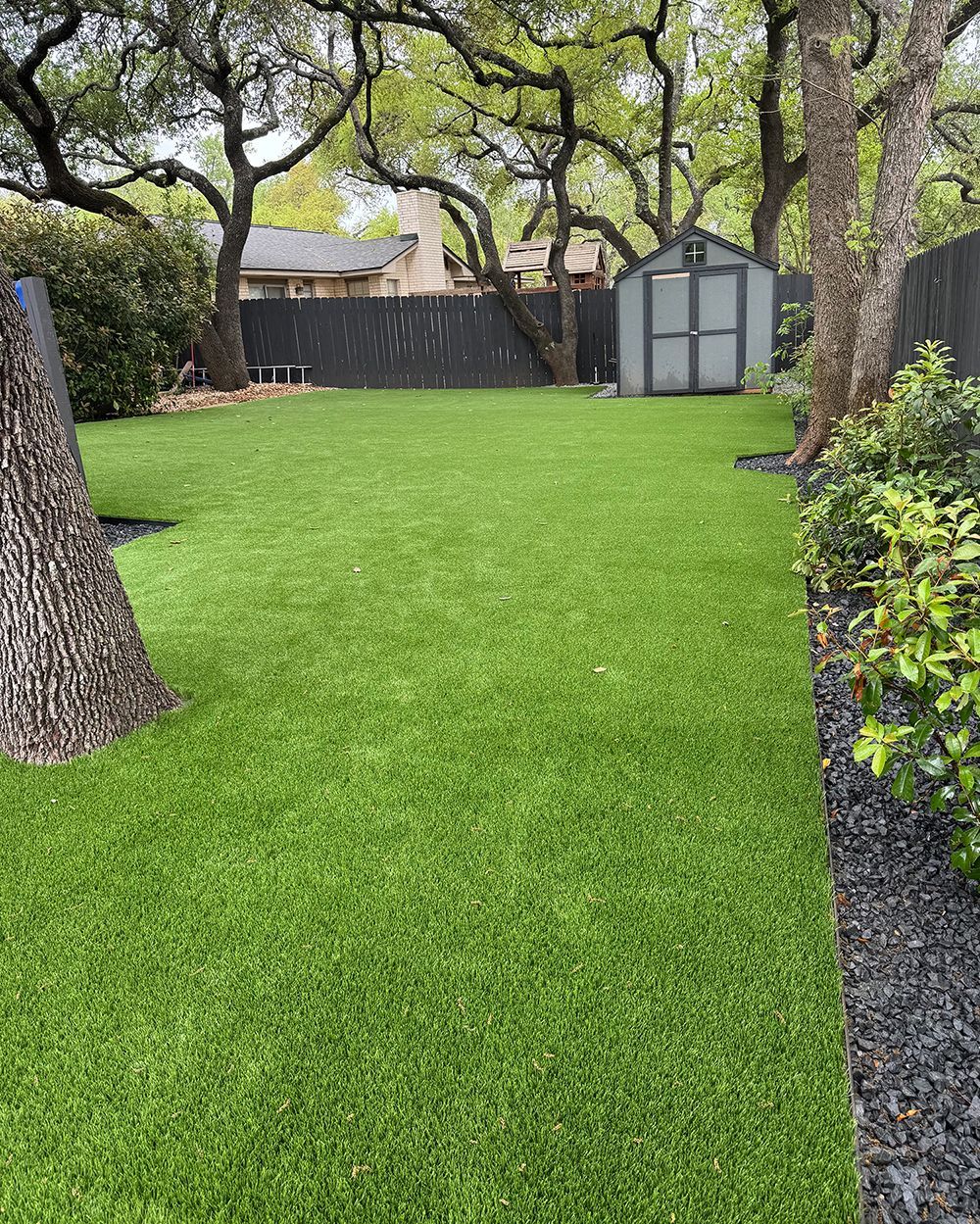 Backyard with artificial green turf, a gray shed, and trees against a dark fence.