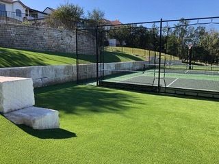 Green turf area with tennis court behind black netting; stone wall and house in the background.